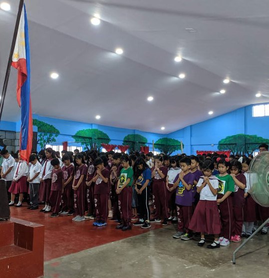St. Mark pupils in maroon and white uniforms standing for the flag ceremony in the school's covered court.